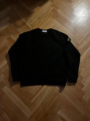 Svart Stone Island sweatshirt - Svart sweatshirt från Stone Island med klassisk rund hals och ribbade muddar. Tröjan har den ikoniska Stone Island-loggan på vänster ärm, broderad i gult och grönt. Materialet är mjuk bomull och passformen är normal. Perfekt för dig som gillar streetwear och snygga detaljer.