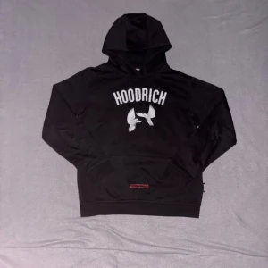 Svart Hoodie från Hoodrich  - Svart hoodie från Hoodrich med stor vit logga framtill och tryckta vita duvor. Baksidan har coolt motiv med duvor och röd text 'From Nothing To Something'. Huva med röd broderad text, känguruficka och mjukt material. Perfekt streetwear för dig som vill sticka ut.