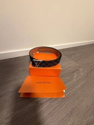 Louis Vuitton rutig skinnbälte - Säljer ett Louis Vuitton bälte i svart och grått med klassiskt rutigt Damier-mönster och en snygg LV-spänne i silver. Bältet är tillverkat i äkta skinn och har en stilren, bred form som ger en lyxig vibe. Kommer med originalförpackning och kvitto.