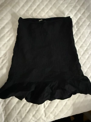 Svart volangkjol från Bik Bok XS - Snygg svart kortkjol från Bik Bok i storlek XS. Kjolen har en enkel design med volangkant nedtill. Materialet känns lätt och funkar både som kjol och topp, men original designen är en kjol!