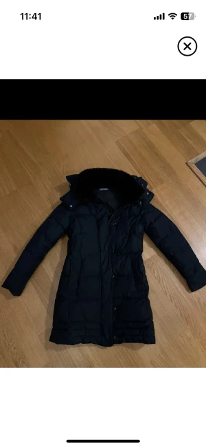 Mörkblå  lång dunjacka Tommy Hilfiger XS - Mörkblå/svart  lång dunjacka från Tommy Hilfiger med snyggt emblem på ärmen och fluffig krage. Jackan har dragkedja och knappar framtill, två fickor och quiltad design. Perfekt för kalla dagar med stilren look och skön passform.