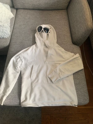 Vit hoodie med goggles från C.P. Company - Säljer en clean vit hoodie från C.P. Company med deras ikoniska goggles i huvan. Tröjan har half zip, stor magficka och justerbar snörning i huvan. Perfekt för dig som vill sticka ut med streetwear-vibe och gillar detaljer. Materialet är mjuk bomull och passformen är normal. Det är en S men funkar både till xs och S. Skicka gärna pris förslag!