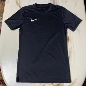 Svart Nike Dri-FIT t-shirt - Svart t-shirt från Nike med klassisk vit swoosh-logga på bröstet. Tillverkad i lätt och funktionell polyester med Dri-FIT-teknologi som håller dig torr. Modellen har rund hals och korta ärmar, perfekt för sport och träning.