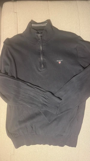 Marinblå GANT half zip tröja M - Marinblå långärmad tröja från GANT i storlek M med half zip och ståkrage med vita ränder på insidan. Tröjan har GANT-logga broderad på bröstet och är tillverkad i mjuk bomull. Ribbstickade muddar vid ärmslut och nederkant ger en clean look. Hör av dig om du har frågor!