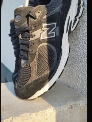 New Balance 2002R Svarta Sneakers - Säljer ett par New Balance 2002R sneakers i svart med grå detaljer. Skorna har meshpaneler och mockaöverdrag, samt den klassiska N-loggan på sidan. De har snörning och en vit, chunky sula för extra komfort. Perfekta för dig som gillar stilrena och sportiga sneakers.