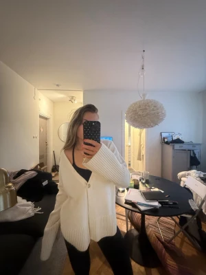 Vit stickad kofta från Busnel - Säljer en oversized vit stickad kofta från Busnel med bred ribbstickning och stora ärmar. Koftan har v-ringning, guldknapp framtill och en snygg logga på ärmen. Perfekt för lager-på-lager och chill dagar.