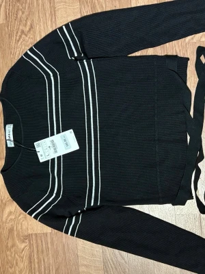 Svart ribbad kofta från Zara - Säljer en svart ribbad kofta från Zara med vita ränder över bröstet och ärmarna. Tröjan har långa ärmar och ett knytband i midjan för extra detalj. Perfekt för dig som gillar stilrena och sportiga vibbar.