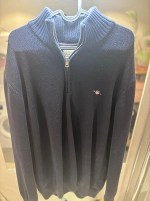 Mörkblå stickad tröja från Gant - Mörkblå stickad tröja från Gant med halv dragkedja och ribbad krage. Klassisk och stilren design med broderad Gant-logga på bröstet. Perfekt för lager-på-lager och passar till många olika outfits.