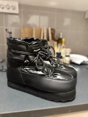 Svarta moon boots från Juicy Couture strl 41 - Säljer ett par svarta moon boots från Juicy Couture med hög platåsula och snörning framtill. Skorna har en glansig finish, broderad Juicy-logga bak och dragsko upptill. Tillverkade i textil och PU med mjukt foder för extra värme. Skorna är endast använda ute vid två tillfällen.                                         på sista bilden kan ni se ett tillverkningsfel där loggan är satt upp och ner men påverkar inte funktionen någonting. Skorna är inhandlade på Boozt och kvitto finns. 