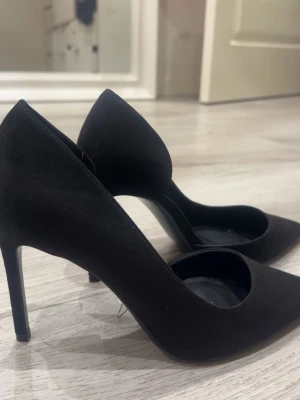 Svarta pumps med spetsig tå - Säljer ett par klassiska svarta pumps i storlek 39 med spetsig tå och hög klack. Skorna har en elegant siluett och är tillverkade i mockaimitation som ger en lyxig känsla. Perfekta för dig som vill ha en stilren och tidlös look.