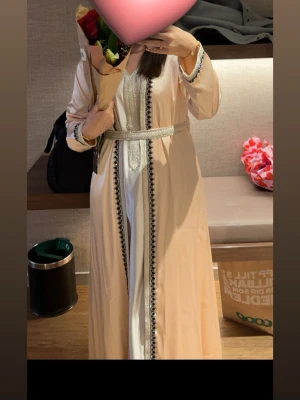 Lång ljus kaftan med silverdetaljer - Säljer en elegant lång kaftan i ljus beige och vitt från D.D Fashion. Klänningen har vackra silverbroderier längs kanterna, svarta dekorativa detaljer och ett matchande bälte i midjan. Materialet är glansigt och ger en lyxig känsla. Perfekt för dig som vill sticka ut. Denna klänning är aldrig använd och köpt för 1225 kr. 