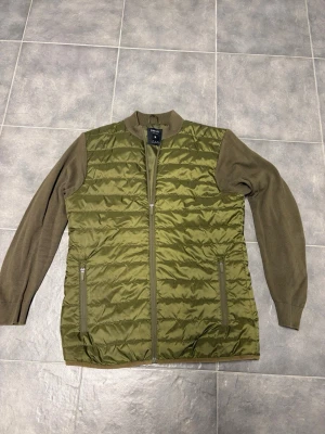 Olivgrön quiltad jacka från SMOG - Säljer en olivgrön quiltad jacka från SMOG i slim fit. Jackan har stickade ärmar och krage, samt två fickor med dragkedja framtill. Kroppen är i glansig polyester med horisontella sömmar och ärmarna är mjukt stickade för extra komfort.