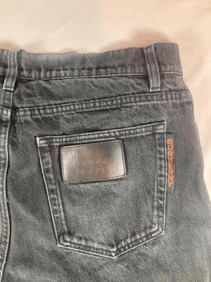 Svarta jeans från Burberry - Säljer ett par svarta jeans från Burberry med klassisk femficksdesign och Burberry-logga på bakfickan samt i midjan. Jeansen har rak passform och är tillverkade i slitstarkt bomullsmaterial. Snygga detaljer med metallknappar och dragkedja framtill. Storlek är W32 och längd 95cm 