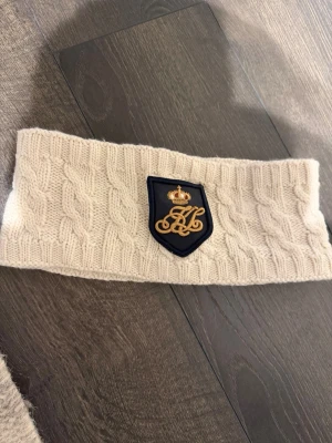 Cremevit stickad pannband med emblem - Säljer ett stickat pannband i färgen cremevit med flätmönster och ett snyggt marinblått emblem med gulddetaljer framtill. Utsidan är i ullblandning och insidan är fodrad med mjuk fleece för extra värme. Perfekt accessoar för kyliga dagar.