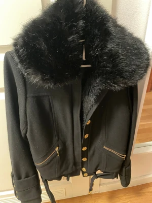 Svart jacka med päls  och guldknappar - Snygg svart jacka med stor krage päls , guldiga knappar och dragkedjor på fickorna. Jackan har bälte i midjan och vid ärmsluten för extra stil. Perfekt för dig som vill ha en statementjacka med lyxig känsla.