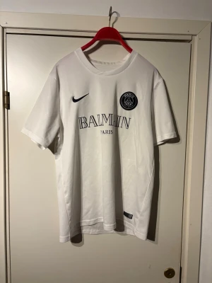 Vit PSG x Balmain t-shirt Nike L - Säljer en clean vit PSG x Balmain t-shirt från Nike i storlek L. T-shirten har korta ärmar, rund hals och tryck med Balmain Paris och PSG-logga på bröstet. Tillverkad i lätt och andningsbar polyester med Dri-FIT-teknologi.