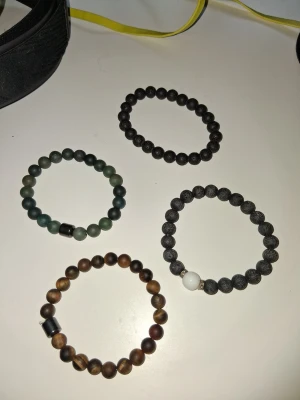 4 STYCKEN ARMBAND  -  Ett armband i mattsvart lavasten, ett i grönmelerad sten, ett i bruna tigeröga-pärlor och ett svart med en vit accentpärla.