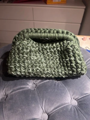 Virkad clutch - Unik handväska i mörkgrönt med lite glitter virkat material med grov struktur och inbyggt handtag. Perfekt för dig som vill ha en cool accessoar som sticker ut. Väskan är mjuk och har en boxig form, vilket gör den både snygg och praktisk för småsaker.