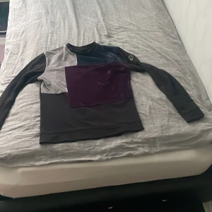 Patchwork sweatshirt från Le Vichy - Unik långärmad sweatshirt från Le Vichy med patchwork-design i mörklila, silver och svart. Tröjan har en cool badge på ärmen och är gjord i mjuk sammet och polyester. Perfekt för dig som gillar statement-plagg och vill sticka ut med din stil.