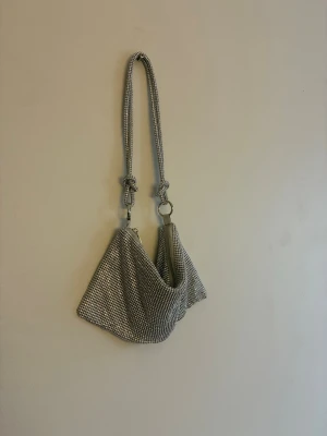 Glittrig silver axelväska - Säljer en glittrig axelväska i silver med massor av små stenar över hela väskan. Väskan har en cool kedjerem med knutdetaljer och stängs med dragkedja. Perfekt för dig som vill sticka ut och addera lite extra bling till din outfit.