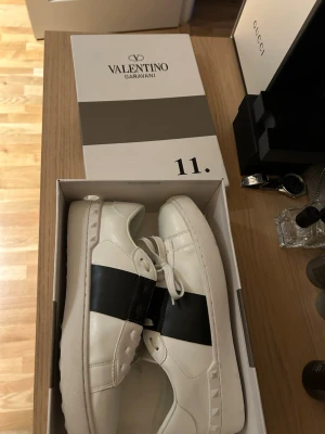 Valentino Garavani Open sneakers vit/svart - Säljer ett par Valentino Garavani Open sneakers i vitt skinn med bred svart läderdetalj över mitten. Klassisk chunky sula med nitar på sidan och platt sula. Snygg och stilren design som sticker ut med den svarta kontrasten.