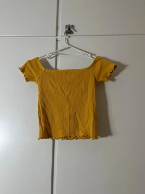 Gul ribbad topp från Stradivarius - Säljer en söt gul ribbad topp från Stradivarius i storlek M. Toppen har korta ärmar, offshoulder-modell och vågig kant nertill och vid ärmslut. Perfekt till sommaren och enkel att styla med jeans eller kjol.