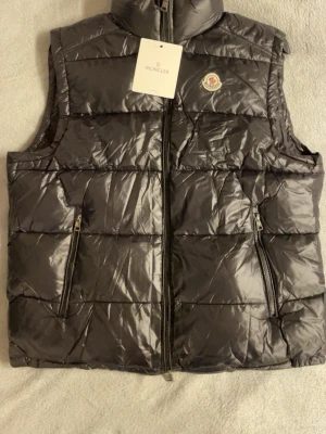 Svart dunväst från Moncler - Snygg svart dunväst från Moncler med glansig finish och klassisk logga på bröstet. Västen har hög krage, två fickor med dragkedja och quiltad design. Perfekt att slänga över en hoodie eller tröja för extra värme.