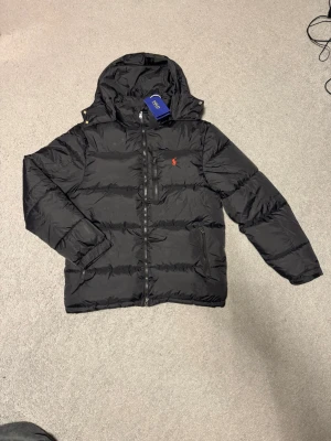 Svart pufferjacka från Polo Ralph Lauren - Säljer en svart pufferjacka från Polo Ralph Lauren i storlek M. Jackan har huva, dragkedja framtill, tre fickor med dragkedja och den klassiska röda Polo-loggan broderad på bröstet. Perfekt för kalla dagar och riktigt snygg till streetwear-stilen. Fråga gärna om du har några frågor