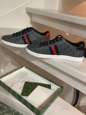 Gucci sneakers med GG-mönster - Säljer ett par stilrena Gucci sneakers i mörkgrått med GG-monogram, klassiska blå och röda ränder på sidan och kontrasterande häl i rött och blått. Skorna har vita sulor, svarta skosnören och är tillverkade i canvas och skinn. Perfekta för dig som vill ha något extra till din outfit. Oanvända lite stora i storleken
