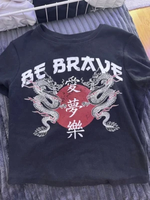 Svart t-shirt med drakmotiv och text - Cool svart t-shirt med tryck av två vita drakar, röd cirkel och asiatiska tecken. Texten 'BE BRAVE' syns över motivet. T-shirten har korta ärmar och en klassisk rund hals. Perfekt för dig som gillar streetstyle och grafiska prints.