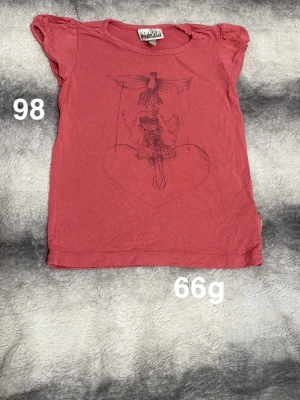 Rosa/röd t-shirt med tryck från Pompdelux - Söt rosa/röd t-shirt från Pompdelux med korta puffärmar och ett tryck av en flicka på en gunga som hålls av en fågel. Perfekt för dig som gillar lekfulla motiv och mjuka material.🌸 Fint använt skjck. Storlek 98 (rosa/röd färg)