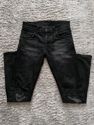 Svarta Nudie Jeans - Säljer ett par svarta jeans från Nudie Jeans med  riktigt snygga slitna detaljer och fetta slitningar på knäna. Jeansen har klassisk femficksdesign, slim fit och diskret broderad logga på bakfickan. Jeansen är i Nudie jeans modellen Grim Tim. Perfekta för dig som gillar en avslappnad och fett stil. W29/32L