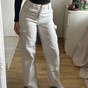 Beiga cargopants med vida ben - Säljer ett par beiga cargopants med vida ben och stora fickor på sidorna. Byxorna har hög midja och stängs med knapp och dragkedja. 