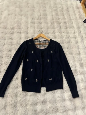 Mörkblå kofta med rosett-detaljer Robin - Mörkblå kofta med dekorativa kaniner detaljer. Koftan har en rund halsringning och knäpps med knappar framtill. Insidan av kragen har ett klassiskt rutmönster. Några av kaninflugorna har lossnat av, se bilder