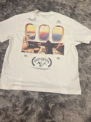Vit t-shirt soft enkel still passar mycket bra till sommaren. Aldrig använd! - Säljer en vit t-shirt från Bershka i storlek S med coolt tryck av flygplansfönster och texten 'Dopamine' på ryggen. Framsidan har tryck med 'Departures' och listade städer. Skön passform och tillverkad i mjuk bomull. Pris kan diskuteras!