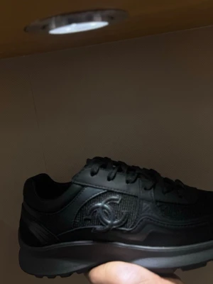 Svarta sneakers från Chanel - Säljer ett par svarta sneakers från Chanel med ikoniska CC-loggan på sidan. Skorna har snörning, rund tå och en chunky sula. Materialmix av skinn och textil ger en clean och lyxig vibe. Perfekta för dig som vill ha en stilren och exklusiv look. Size 42/43