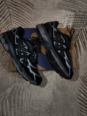 Svarta och grå Asics sneakers - Säljer ett par snygga Asics sneakers i svart och grått med vita detaljer. Skorna har meshpaneler för bra ventilation, klassisk snörning och en robust sula som ger grym komfort. Perfekta för dig som gillar sportig stil och vill ha något som funkar till jeans eller träningskläder.