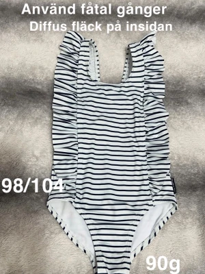 Randig baddräkt med volanger - Säljer en söt baddräkt i vitt med marinblå ränder och volangdetaljer längs axelbanden. Perfekt för strand eller pool och har en klassisk, stilren look. 🌸 Använd fåtal gånger. En diffus fläck på insidan ( ses inte utifrån). Storlek 98/104