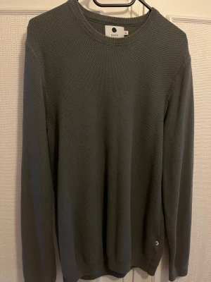Mörkgrå stickad tröja från NN07 - Säljer en mörkgrå stickad tröja från NN07 i storlek XL. Tröjan har en rund halsringning, diskret strukturmönster och långa ärmar. Perfekt för dig som gillar enkel och stilren design. Passar till både jeans och chinos.