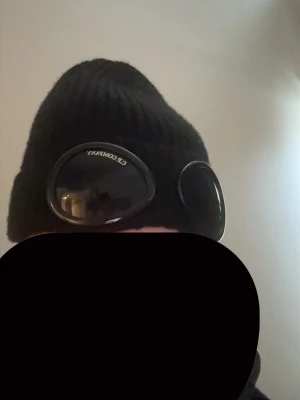 Svart mössa från C.P. Company - Säljer en svart ribbstickad mössa från C.P. Company med ikoniska goggles framtill. Mössan är i mjukt material och har en clean look som passar perfekt till streetwear-stilen. Märket syns tydligt på linsen och insidan har äkthetsmärkning.