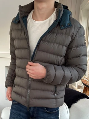 Grå pufferjacka från Colmar - Säljer en grå pufferjacka från Colmar Originals med huva och dragkedja framtill. Jackan har en stilren look med horisontella sömmar och två fickor med dragkedja. Perfekt för kyliga dagar och riktigt skön att bära.