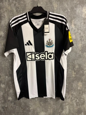 Newcastle United Isak fotbollströja - Large - Säljer en svartvit randig Newcastle United matchtröja från Adidas med Isak 14 på ryggen. Tröjan har klassiska tre ränder på axlarna, klubbmärke på bröstet och sponsorlogga framtill. Tillverkad i lätt polyester, perfekt för fotboll eller som supporterplagg.