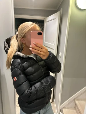 Svart pufferjacka från Colmar - Säljer en svart pufferjacka från Colmar med huva och dragkedja framtill. Jackan har en klassisk quiltad design och en rund Colmar-logga på ärmen. Perfekt för kyliga dagar och enkel att matcha med olika outfits.