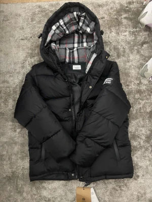 Svart pufferjacka från Burberry M - Säljer en svart pufferjacka från Burberry med stor logga på ärmen och rutigt foder i huvan. Jackan har dragkedja, knappar och en mysig, vadderad känsla. Perfekt för kalla dagar och riktigt snygg streetstil.