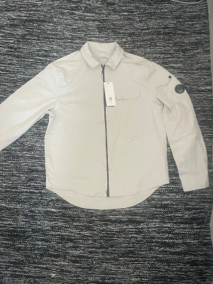 Beige overshirt från C.P. Company - Aldrig använd då den är för liten för mig. Snygg beige overshirt från C.P. Company med klassisk krage, svart dragkedja och har en bröstficka med lock, jackan är tillverkad i ett lätt syntetmaterial som passar perfekt för lager-på-lager. Perfekt för dig som gillar stilrena och moderna plagg. Rep1:1