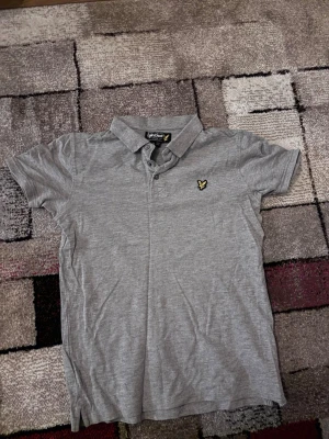 Grå piké från Lyle & Scott Junior - Säljer en stilren grå pikétröja från Lyle & Scott Junior med klassisk krage, två knappar och den ikoniska gula loggan på bröstet. Perfekt till jeans eller shorts för en clean look.