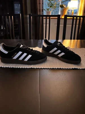 Adidas Spezial svarta sneakers mock soppa - Säljer ett par Adidas Spezial sneakers i svart mocka med klassiska vita ränder och detaljer. Skorna har brun sula, snörning och guldfärgad Spezial-logga på sidan. Perfekta för dig som gillar retrostil och streetwear.