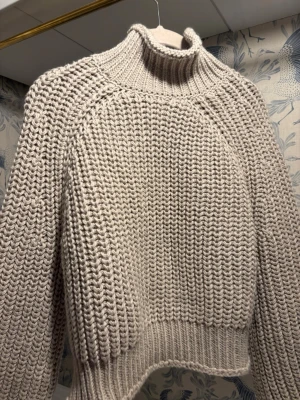 Beige chunky stickad polotröja - Chunky stickad polotröja i beige med bred ribbad krage och muddar. Långa ärmar och grovstickad struktur som ger en riktigt mysig vibe. Perfekt för kyliga dagar när du vill vara både varm och snygg.