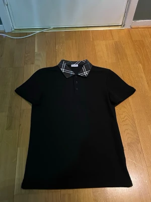 Svart Burberry piké med rutig krage - Säljer en svart pikétröja från Burberry med klassisk rutig krage i svart, vitt och blått. Tröjan har korta ärmar och tre knappar framtill. Materialet känns som bomull och modellen är normal i passformen. Perfekt för dig som gillar stilrena plagg med en twist.