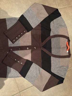 Randig kofta från 365 Sunshine, M - Snygg kofta med breda ränder i brunt, grått, beige och svart från 365 Sunshine. Långärmad modell med knappar framtill och vid ärmsluten. Mjuk och skön i stickad bomullsmix, perfekt för lager på lager under höst och vinter. Liten i storlek, en knapp saknar (se bild). 